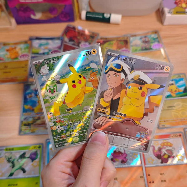 Dos cartas Pokémon brillantes en chino con Pikachu, sostenidas sobre una mesa llena de cartas coleccionables.